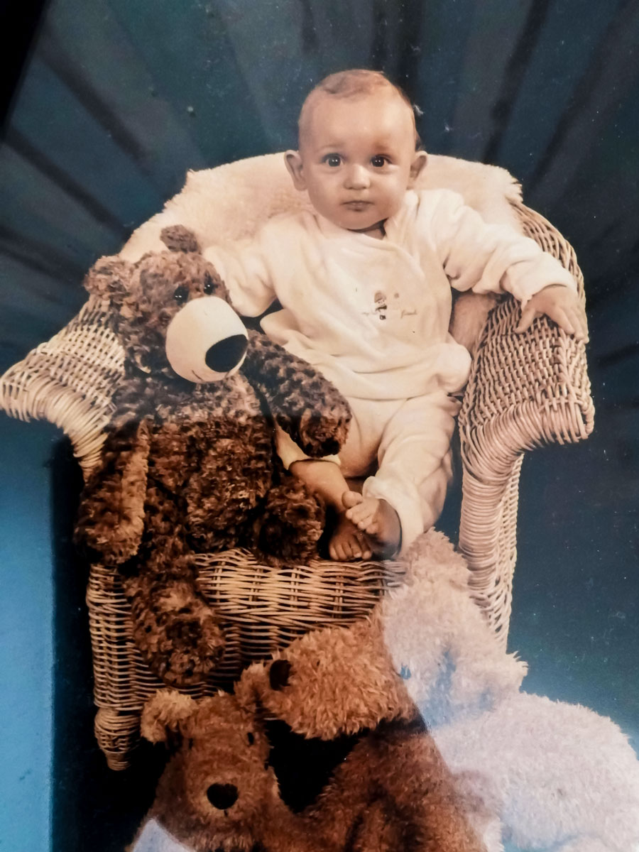 Kinderfoto von Julian, der auf einem Stuhl sitzt. Vor ihm auf dem Stuhl sitzt ein Stoffbär, und rechts vor dem Stuhl sitzt ein Stoffhund.