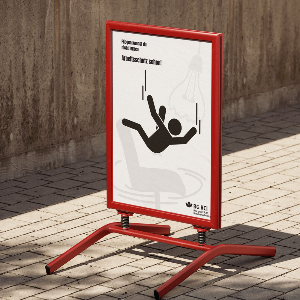 Straßenschild-Mockup mit einem Plakat: Rechts eine Piktogramm-Person im Fall, links daneben ein großer illustrierter Drehstuhl. Oben links steht der Text „Fliegen kannst du nicht lernen. Arbeitsschutz schon.