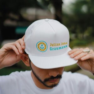 Ein Mützen Mockup mt einen Kombiniertem Logo mit dem Worzeichen Julian John Graumann. Links danben ist das Bildzeichen mit einer Blende und eienr Person mit Schal und Barett-Mütze.